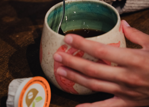 Person holding a ceramic mug with a spoon, next to a tea bag on a wooden surface
