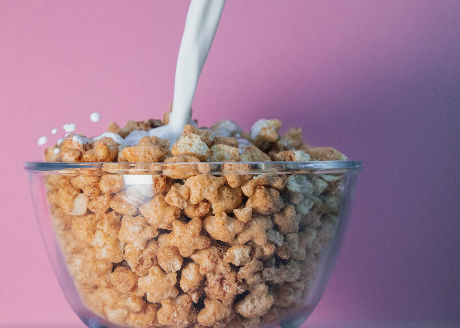 Glass bowl of cereal with milk being poured over it against a pink background