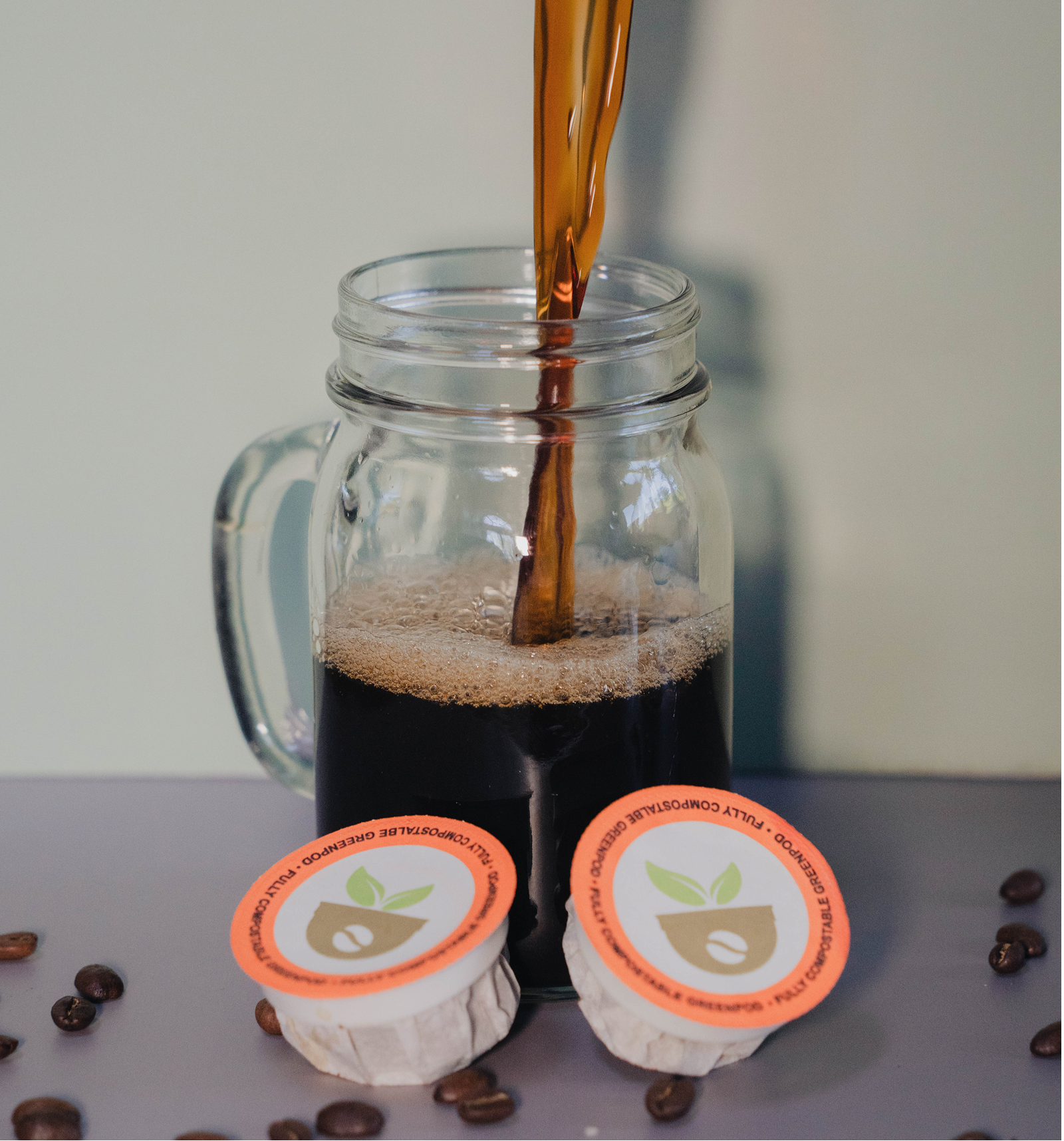 Coffee being poured into a glass mug with coffee pods and beans on a gray surface.