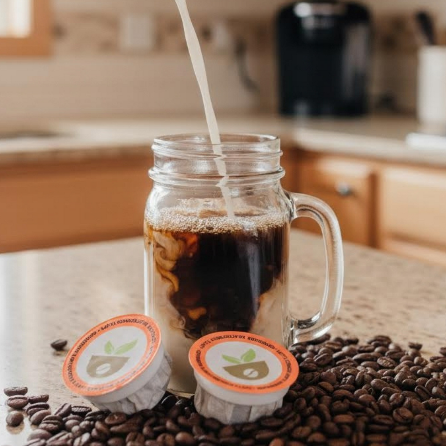 Milk being poured into a glass of coffee with coffee pods and beans on a kitchen counter.