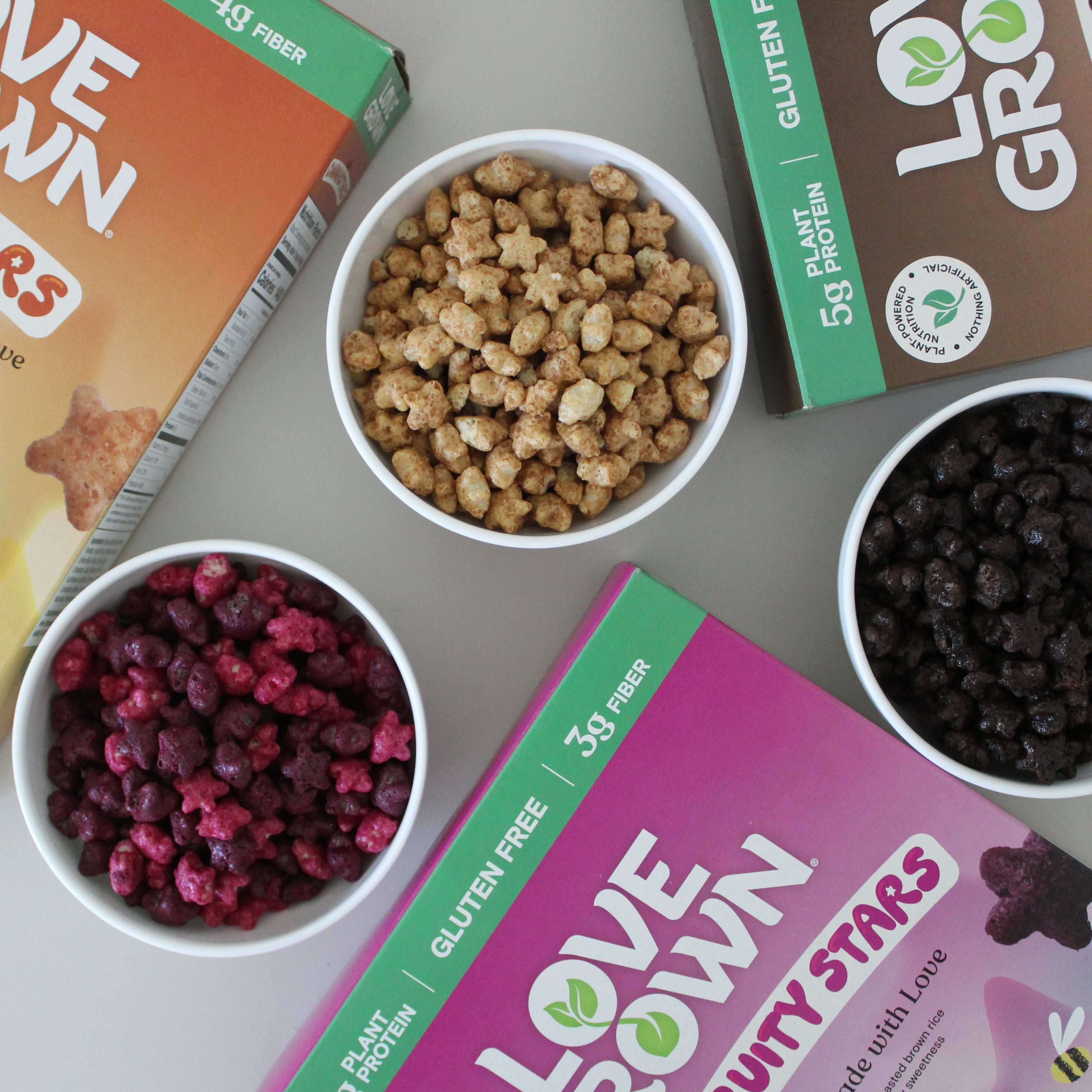 Love Grown Cinnastars, Chocostars and Fruity Stars cereal boxes with bowls of cereal ingredients on a gray surface.