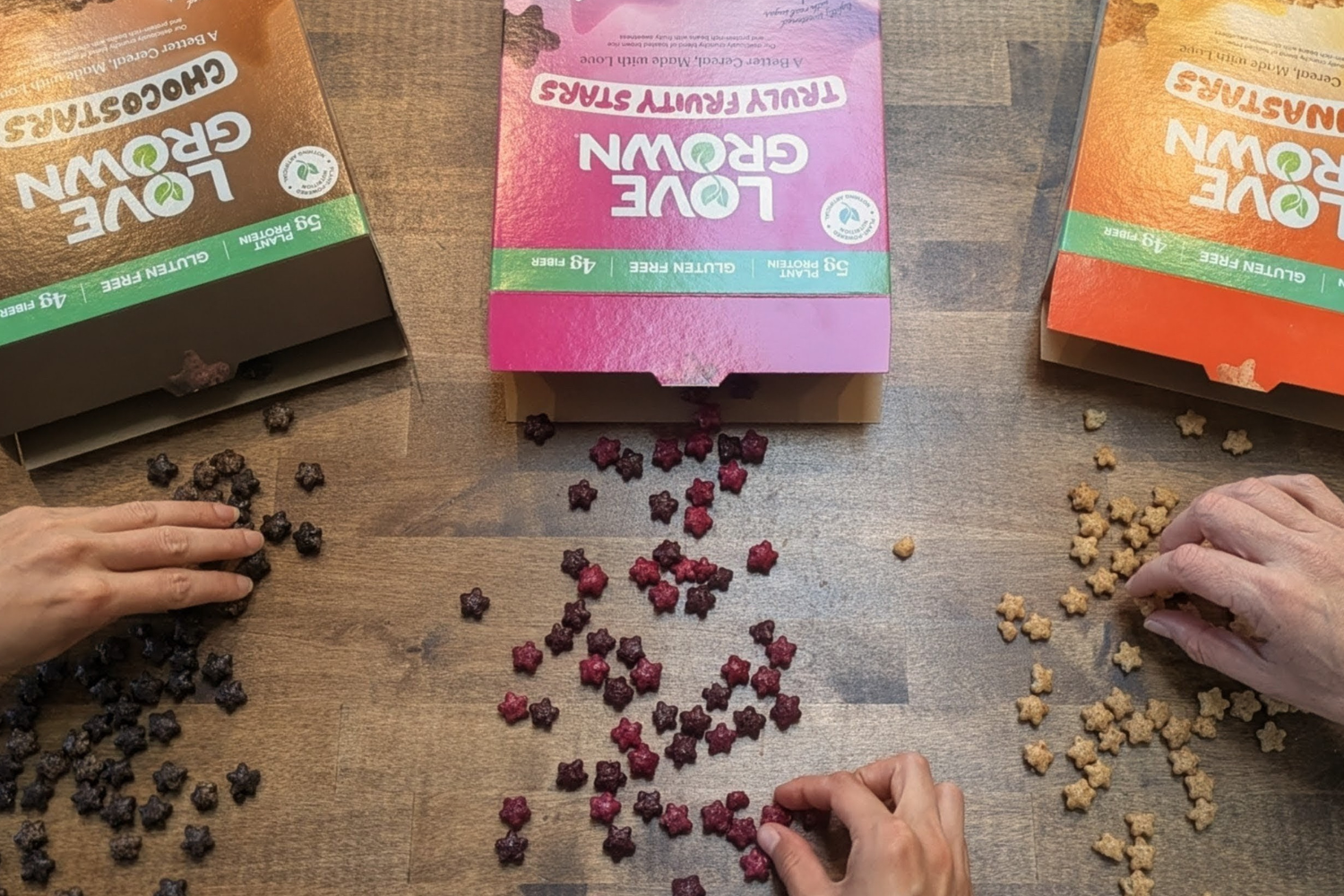 Hands grabbing Love Grown cereal on a wooden surface with Love Grown Food packaging.