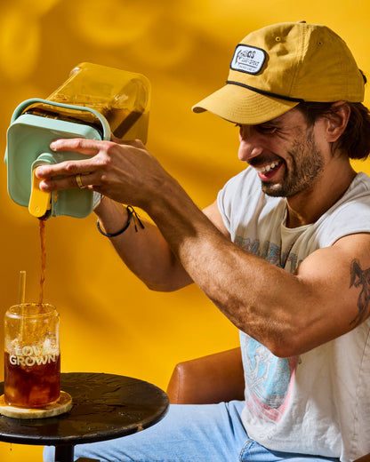 A man wearing a yellow cap and white t-shirt sits in a brown leather chair, pouring dark coffee from a mint green and yellow insulated carafe into an iced drink on a small black table against a bright yellow background.