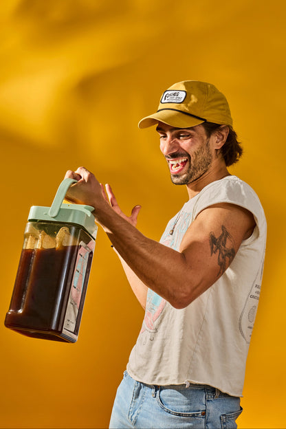 A man wearing a yellow cap and a white sleeveless shirt holds up a large, clear plastic pitcher filled with dark brown liquid, likely beer, with a mint green handle and lid. He is posed against a vibrant, solid yellow background.