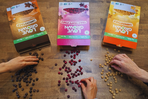 Three boxes of Love Grown Foods on a wooden surface with hands interacting with dried fruits and grains.