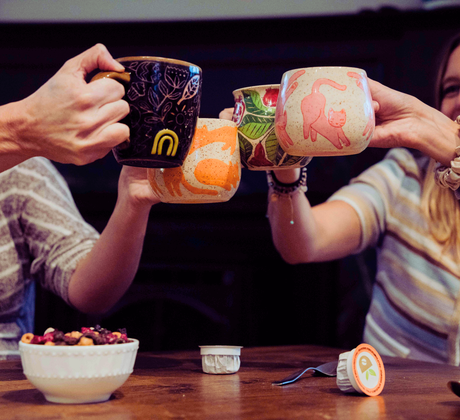 Two people clinking mugs together at a table with snacks and coffee cups.